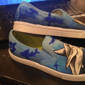 May include: A pair of white canvas slip-on shoes hand-painted with a blue ocean scene. The shoes feature a large shark, smaller sharks, and a blue and white ocean background.