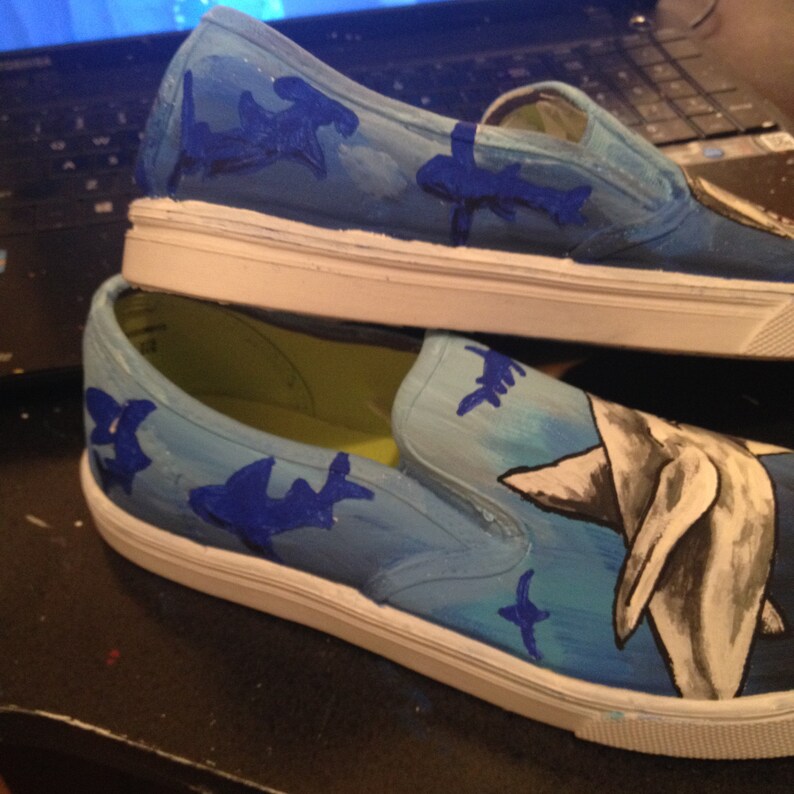 May include: A pair of white canvas slip-on shoes hand-painted with a blue ocean scene. The shoes feature a large shark, smaller sharks, and a blue and white ocean background.