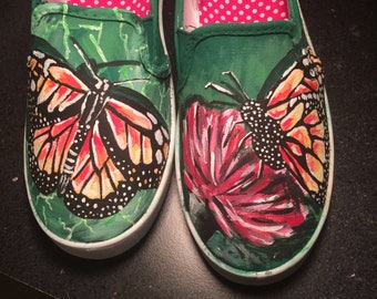 Könnte beinhalten: Ein Paar grüne Canvas-Slipper mit einem bemalten Design aus Schmetterlingen und Blumen. Die Schmetterlinge sind orange, schwarz und weiß, und die Blumen sind rosa und weiß.