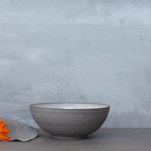 K&ouml;nnte beinhalten: Eine graue Keramikschale mit wei&szlig;em Innenraum steht auf einer grauen Oberfl&auml;che. Ein graues Leinentuch und eine orangefarbene Blume befinden sich links von der Schale. Der Hintergrund ist eine hellgraue Wand.