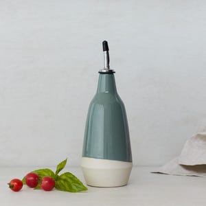 Op de afbeelding: Een keramische olie- en azijnfles met een wit en grijs glazuur. De fles heeft een tuit en een zwarte dop.