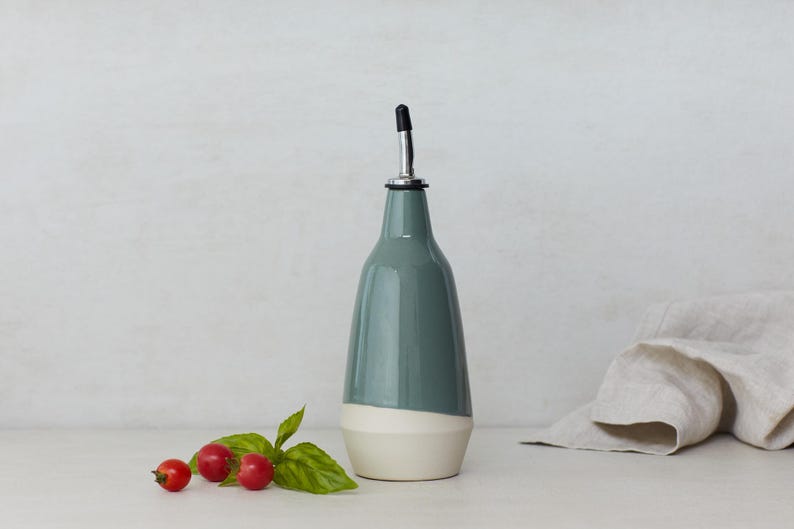 Op de afbeelding: Een keramische olie- en azijnfles met een wit en grijs glazuur. De fles heeft een tuit en een zwarte dop.