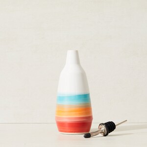 May include: A white ceramic bottle with a colourful, horizontal striped design in shades of red, orange, blue, and white. A stainless steel and black bottle pourer is next to the bottle. The bottle is likely for oil or vinegar.