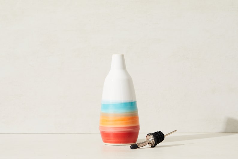 May include: A white ceramic bottle with a colourful, horizontal striped design in shades of red, orange, blue, and white. A stainless steel and black bottle pourer is next to the bottle. The bottle is likely for oil or vinegar.