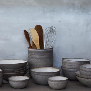 K&ouml;nnte beinhalten: Eine Sammlung grauer Keramik-K&uuml;chenutensilien, darunter Schalen in verschiedenen Gr&ouml;&szlig;en und ein Utensilienhalter mit Holzl&ouml;ffeln und einem Schneebesen. Die Schalen haben eine strukturierte Oberfl&auml;che und eine hellfarbige Innenseite.