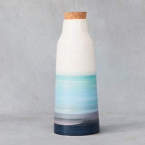 May include: A ceramic bottle with a cork stopper. The bottle is white at the top, transitioning to shades of blue and purple, and ending with a dark blue base. The design resembles a landscape. The bottle is approximately 25 cm tall.