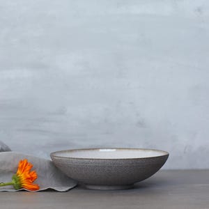 K&ouml;nnte beinhalten: Eine graue Keramikschale mit wei&szlig;em Innenraum steht auf einer grauen Oberfl&auml;che. Ein graues Leinentuch und eine orangefarbene Blume befinden sich links von der Schale. Der Hintergrund ist eine hellgraue Wand.