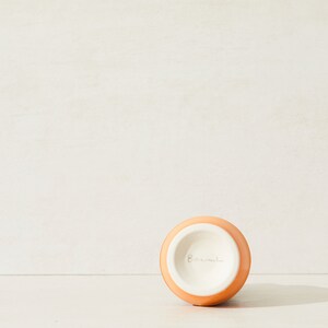 May include: A small, round, orange ceramic vase with a white interior. The word "Barnul" is written in cursive on the bottom of the vase. The vase is set against a neutral background.