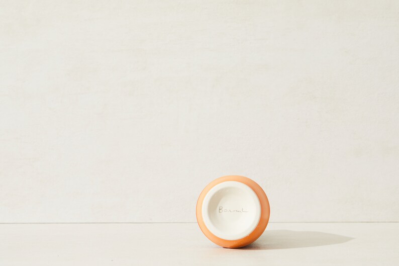 May include: A small, round, orange ceramic vase with a white interior. The word "Barnul" is written in cursive on the bottom of the vase. The vase is set against a neutral background.