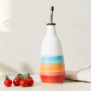 May include: A white ceramic oil bottle with a colourful striped design in shades of red, orange, turquoise, and white. The bottle has a stainless steel spout and is next to a cluster of red cherry tomatoes.
