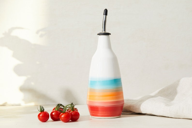 May include: A white ceramic oil bottle with a colourful striped design in shades of red, orange, turquoise, and white. The bottle has a stainless steel spout and is next to a cluster of red cherry tomatoes.