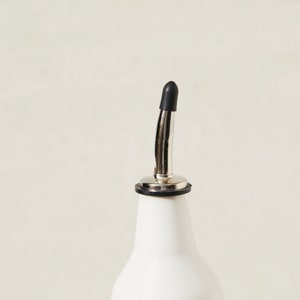 May include: A white ceramic bottle with a stainless steel spout and a black rubber stopper. The bottle is designed for dispensing liquids, such as oil or vinegar. The spout is curved and has a small opening for controlled pouring.