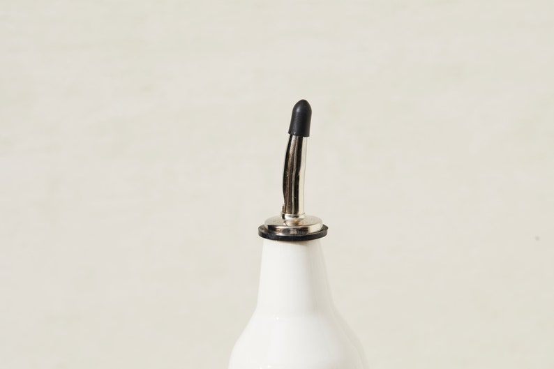 May include: A white ceramic bottle with a stainless steel spout and a black rubber stopper. The bottle is designed for dispensing liquids, such as oil or vinegar. The spout is curved and has a small opening for controlled pouring.