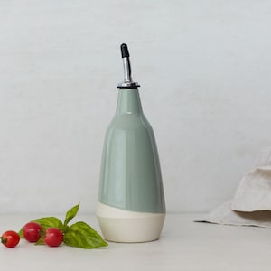 Op de afbeelding: Een keramische olijfolie fles met een witte en lichtblauwe glazuur. De fles heeft een tuit met een zilveren bovenkant en een zwarte gieter. De fles staat op een wit oppervlak met een linnen doek en wat rode bessen.