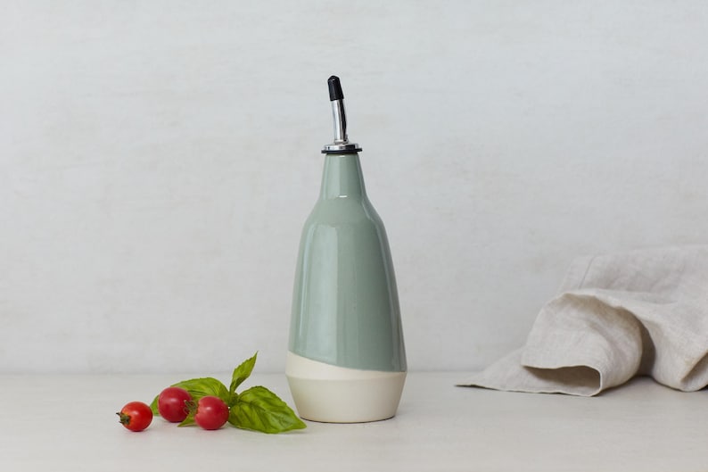 Op de afbeelding: Een keramische olijfolie fles met een witte en lichtblauwe glazuur. De fles heeft een tuit met een zilveren bovenkant en een zwarte gieter. De fles staat op een wit oppervlak met een linnen doek en wat rode bessen.