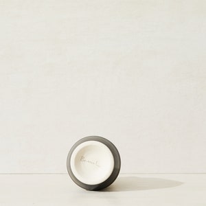 May include: A dark gray ceramic vase with a white interior. The vase has a round base and a narrow opening. The bottom of the vase is marked with the word "Bermel."