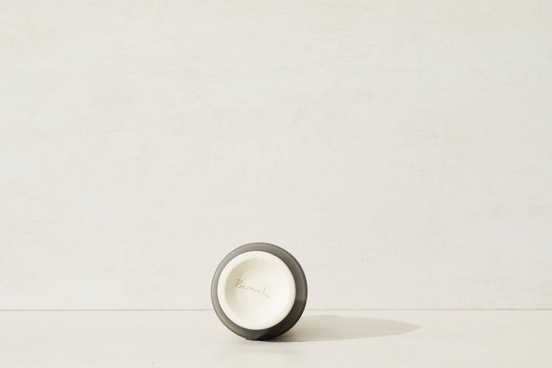 May include: A dark gray ceramic vase with a white interior. The vase has a round base and a narrow opening. The bottom of the vase is marked with the word "Bermel."