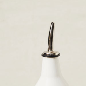 May include: A white ceramic bottle with a stainless steel spout. The spout has a curved design and a black rubber ring at the base. The bottle is a kitchen or barware item.