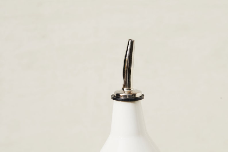 May include: A white ceramic bottle with a stainless steel spout. The spout has a curved design and a black rubber ring at the base. The bottle is a kitchen or barware item.