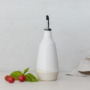 Op de afbeelding: Witte keramische olie- en azijnfles met een zwarte giettuit en een beige bodem. De fles staat op een wit oppervlak met een takje basilicum en drie rode cherrytomaatjes.