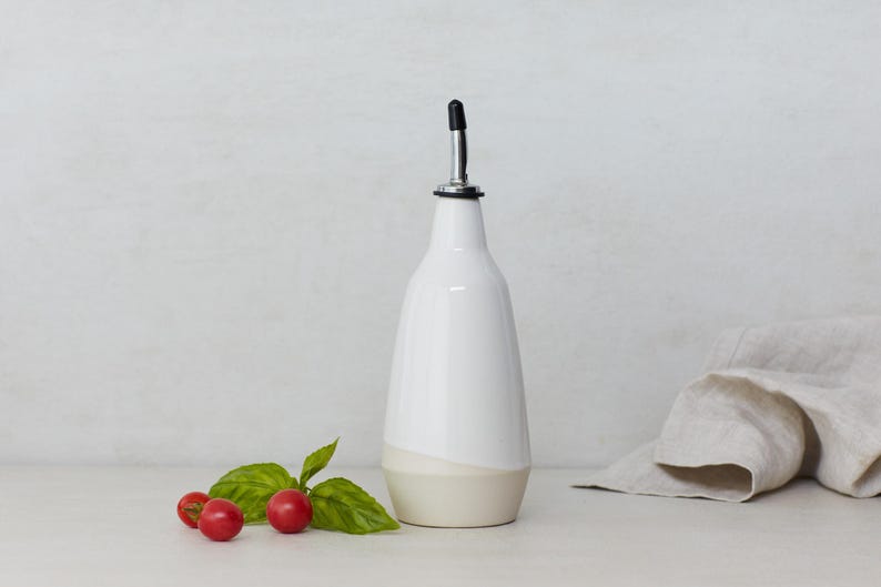 Op de afbeelding: Witte keramische olie- en azijnfles met een zwarte giettuit en een beige bodem. De fles staat op een wit oppervlak met een takje basilicum en drie rode cherrytomaatjes.