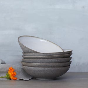 K&ouml;nnte beinhalten: Ein Stapel von sechs grauen Keramikschalen mit wei&szlig;en Innenr&auml;umen. Die Schalen haben eine strukturierte Au&szlig;enseite und ein schlichtes, modernes Design. Ein graues Leinentuch und eine orangefarbene Blume sind im Vordergrund.