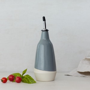 Op de afbeelding: Een grijze keramische fles voor olie en azijn met een witte band rond de bodem. De fles heeft een zilveren tuit en een zwarte dop.