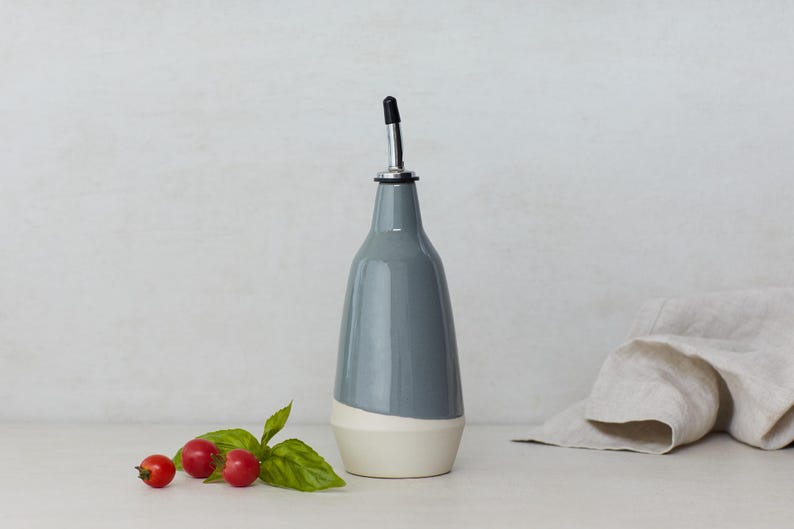Op de afbeelding: Een grijze keramische fles voor olie en azijn met een witte band rond de bodem. De fles heeft een zilveren tuit en een zwarte dop.