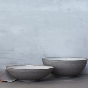 K&ouml;nnte beinhalten: Zwei graue Keramikschalen mit wei&szlig;em Innenraum werden auf einer grauen Oberfl&auml;che pr&auml;sentiert. Die Schalen haben eine strukturierte Au&szlig;enseite und ein schlichtes, modernes Design. Eine orangefarbene Blume und ein graues Tuch befinden sich im Vordergrund.