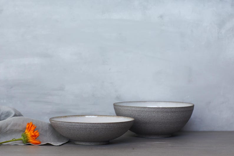 K&ouml;nnte beinhalten: Zwei graue Keramikschalen mit wei&szlig;em Innenraum werden auf einer grauen Oberfl&auml;che pr&auml;sentiert. Die Schalen haben eine strukturierte Au&szlig;enseite und ein schlichtes, modernes Design. Eine orangefarbene Blume und ein graues Tuch befinden sich im Vordergrund.
