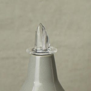 Op de afbeelding: Een glazen flesstopper van helder glas met een puntige bovenkant. De stopper is ontworpen om te passen op een fles met een smalle opening.