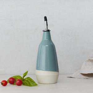 Op de afbeelding: Een keramische olie- en azijnfles met een blauwe en witte glazuur. De fles heeft een tuit met een zwarte dop.