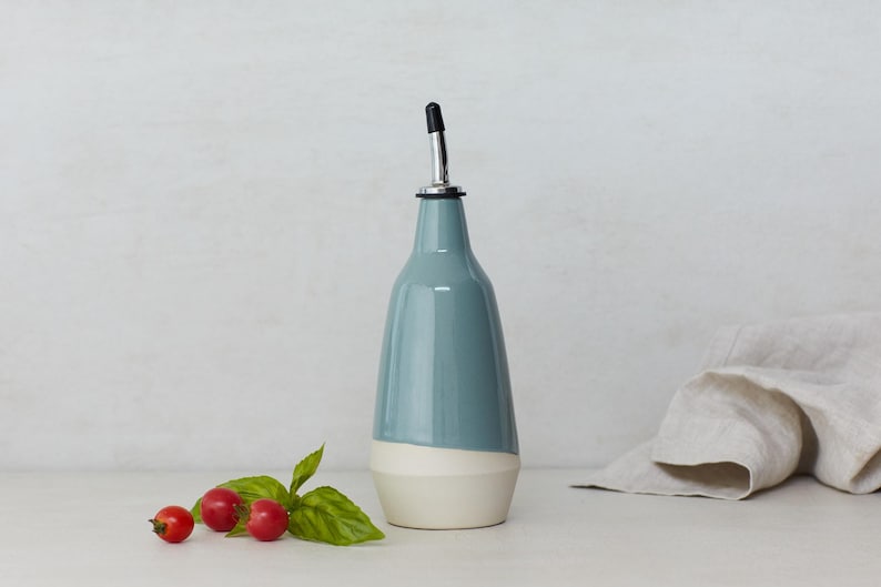 Op de afbeelding: Een keramische olie- en azijnfles met een blauwe en witte glazuur. De fles heeft een tuit met een zwarte dop.