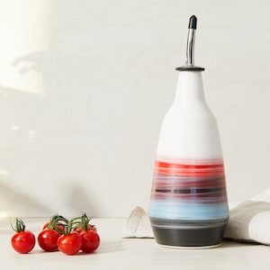 May include: A white ceramic oil and vinegar bottle with a stainless steel pour spout. The bottle has a colourful ombre design with red, pink, blue, and grey stripes. The bottle is sitting on a white surface next to a small group of red cherry tomatoes.