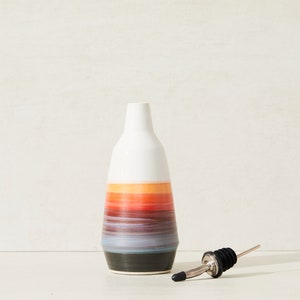 May include: A white ceramic oil and vinegar bottle with a colorful striped design in shades of orange, red, blue, and black. The bottle has a black and silver pour spout.