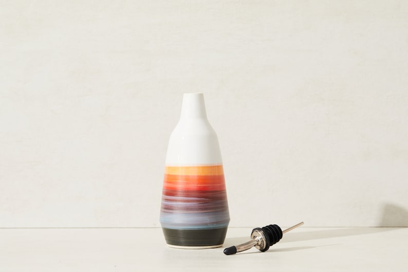 May include: A white ceramic oil and vinegar bottle with a colorful striped design in shades of orange, red, blue, and black. The bottle has a black and silver pour spout.