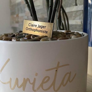 Peut inclure: Un pot en céramique blanche avec le mot "Kurrieta" écrit en lettres dorées. Le pot contient de petites pierres brunes et blanches et une étiquette dorée qui indique "Claire Jager, Orthophoniste".
