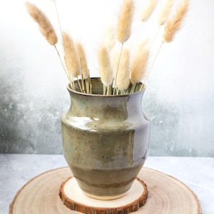 May include: A ceramic vase in shades of green and brown holds dried, fluffy beige flowers. The vase sits on a wooden slice, creating a natural, earthy aesthetic. The arrangement is ideal for home decor.