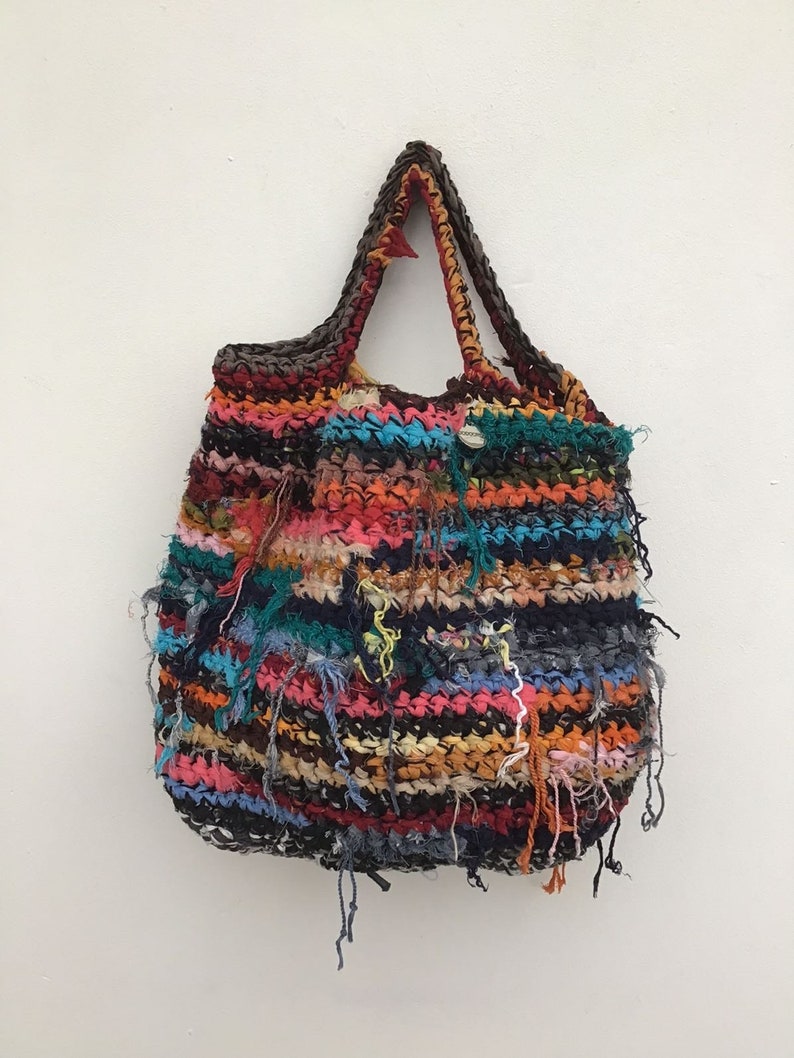 May include: A handmade tote bag with a colorful, textured design. The bag features a variety of colors, including red, blue, orange, and black, with frayed edges. The handles are a dark brown color.