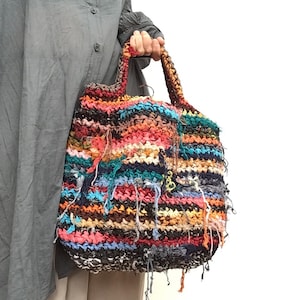 May include: A colorful, handmade tote bag with a textured, woven design. The bag features a mix of vibrant colors including red, blue, orange, and black. The handles are braided, and the bag has fringe details.