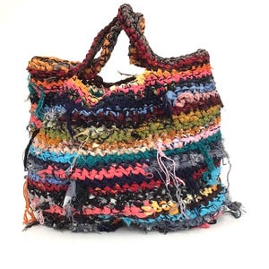 May include: A handmade tote bag with a colorful, textured design. The bag features a variety of colors, including pink, blue, yellow, and black, woven together. The handles are a braided brown and black. The bag has a unique, fringed edge.
