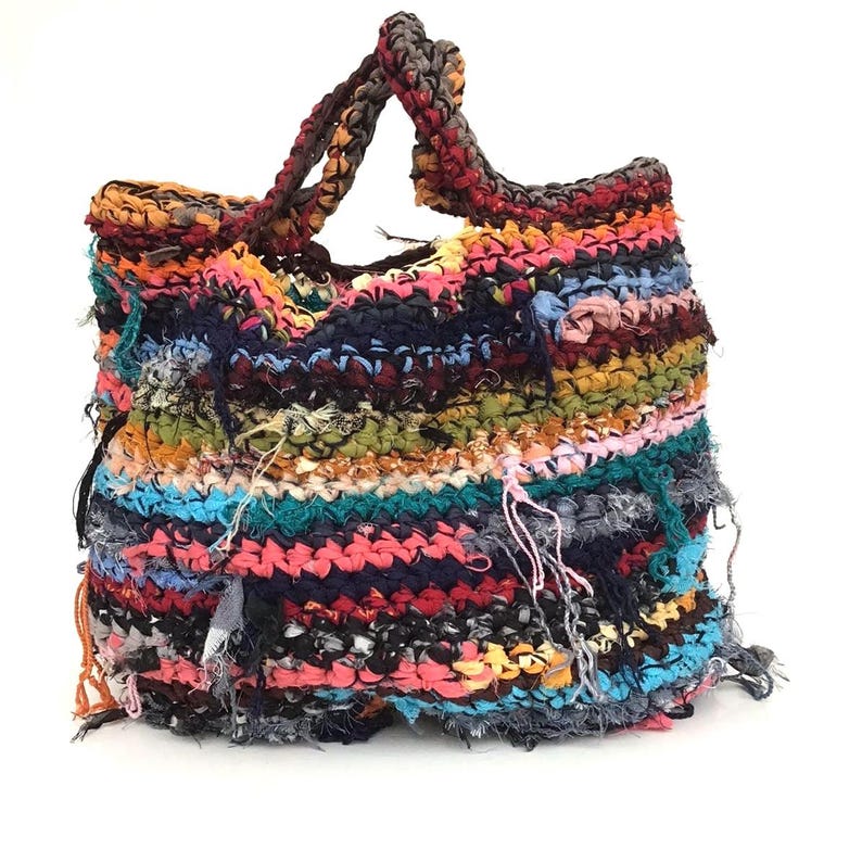May include: A handmade tote bag with a colorful, textured design. The bag features a variety of colors, including pink, blue, yellow, and black, woven together. The handles are a braided brown and black. The bag has a unique, fringed edge.