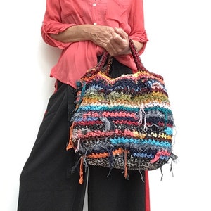 May include: A colorful, handmade tote bag with a textured, woven design. The bag features a mix of blue, orange, green, and black threads, with frayed edges. The handles are a darker, braided material. A unique, artistic accessory.