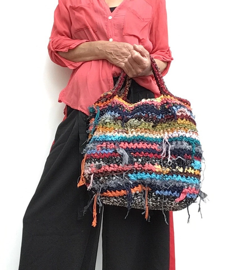 May include: A colorful, handmade tote bag with a textured, woven design. The bag features a mix of blue, orange, green, and black threads, with frayed edges. The handles are a darker, braided material. A unique, artistic accessory.