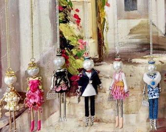 May include: A collection of eleven decorative doll pendants hanging from thin chains. Each doll features a round white bead head and a unique outfit made of fabric, beads, and metallic accents, including dresses, suits, and skirts in various colors.