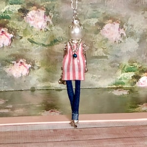 Puede incluir: Un collar de cadena plateada con un dije de vestido a rayas rosa y blanco y jeans azul denim. El dije es una pequeña figura de muñeca con cabeza de perla.