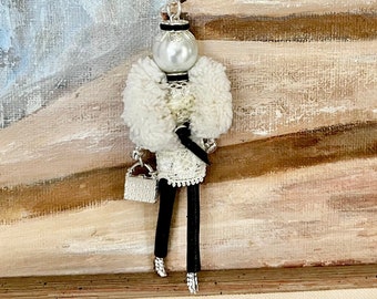 Op de afbeelding: Een witte en zwarte pop met een witte bontjas en een parelketting. De pop houdt een klein wit handtasje vast en staat op een houten oppervlak.