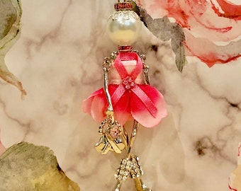 May include: A silver necklace with a pink and white ballerina charm. The ballerina is wearing a pink dress with a white ribbon and a pink flower. The charm is attached to a silver chain with a silver clasp.