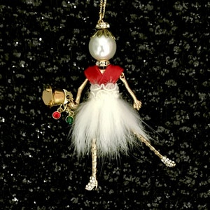 May include: A gold necklace with a white pearl and a red and white ballerina pendant. The ballerina is wearing a white tutu and has a gold bell in her hand.