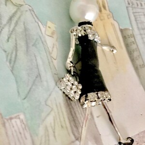 May include: A silver necklace with a pearl and a black and silver figurine of a woman wearing a dress and carrying a purse. The figurine is adorned with rhinestones.